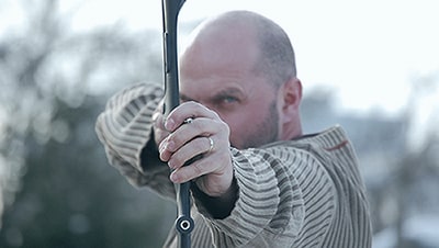 An archer holding a recurve bown dawn aiming at the viewer.